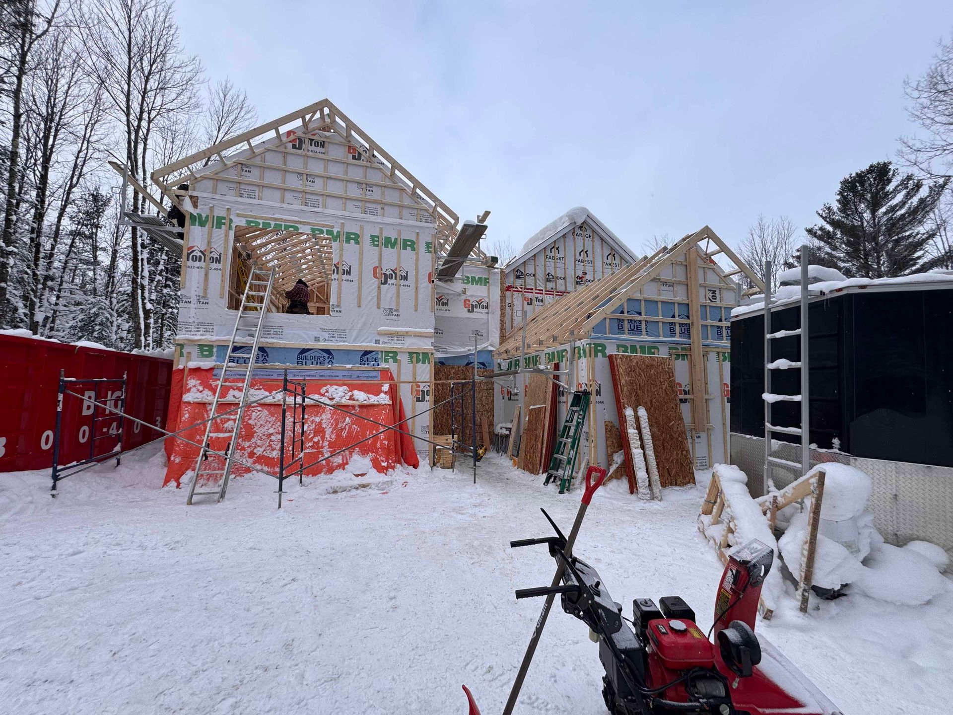 Maison en construction dans un décor enneigé; ossature de bois, pellicule bleue, bâche rouge, souffleuse à neige au premier plan.
