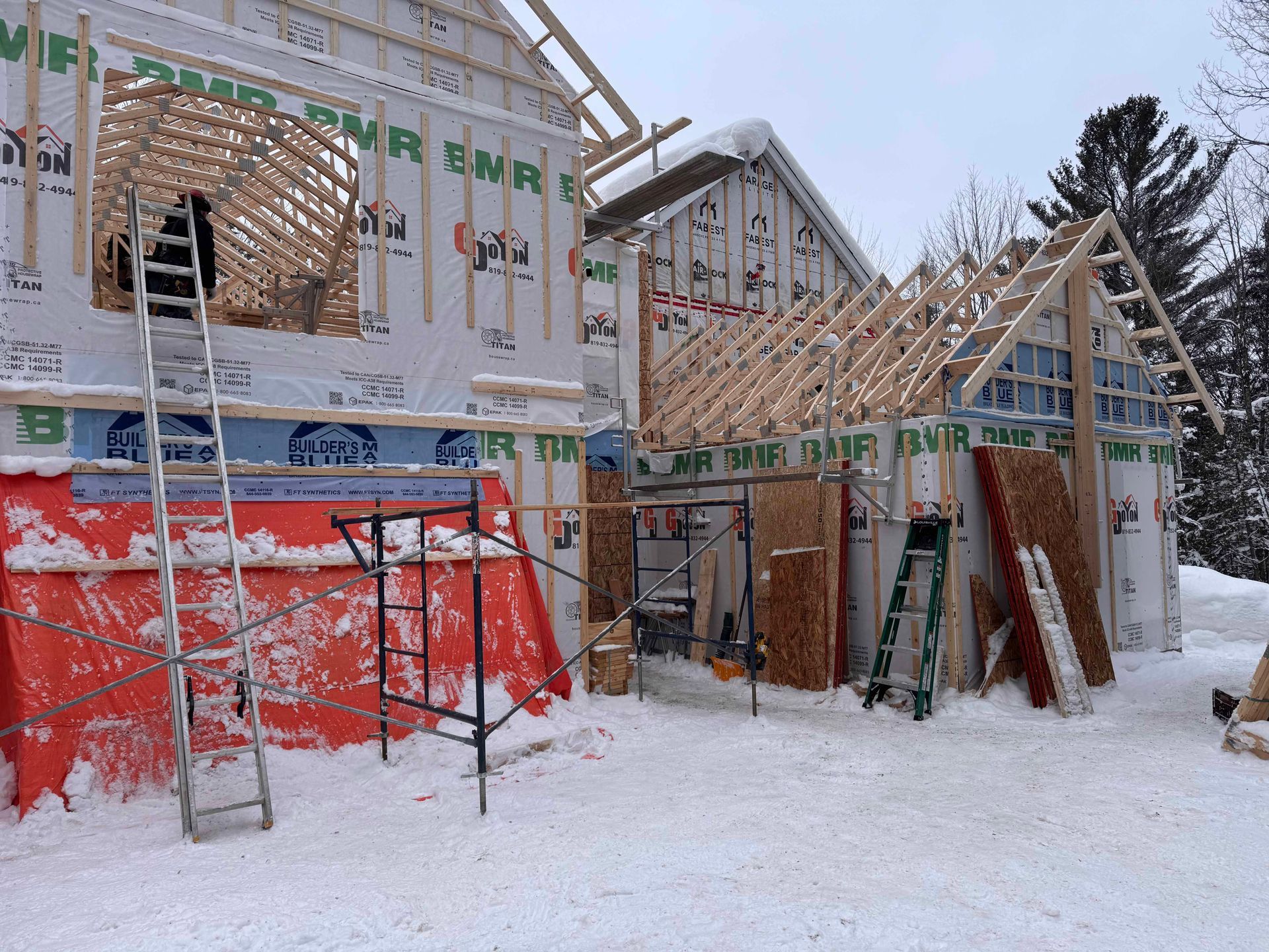 Chantier de construction avec ossature et bardage en bois, recouvert de neige. Une personne sur une échelle, en hiver.