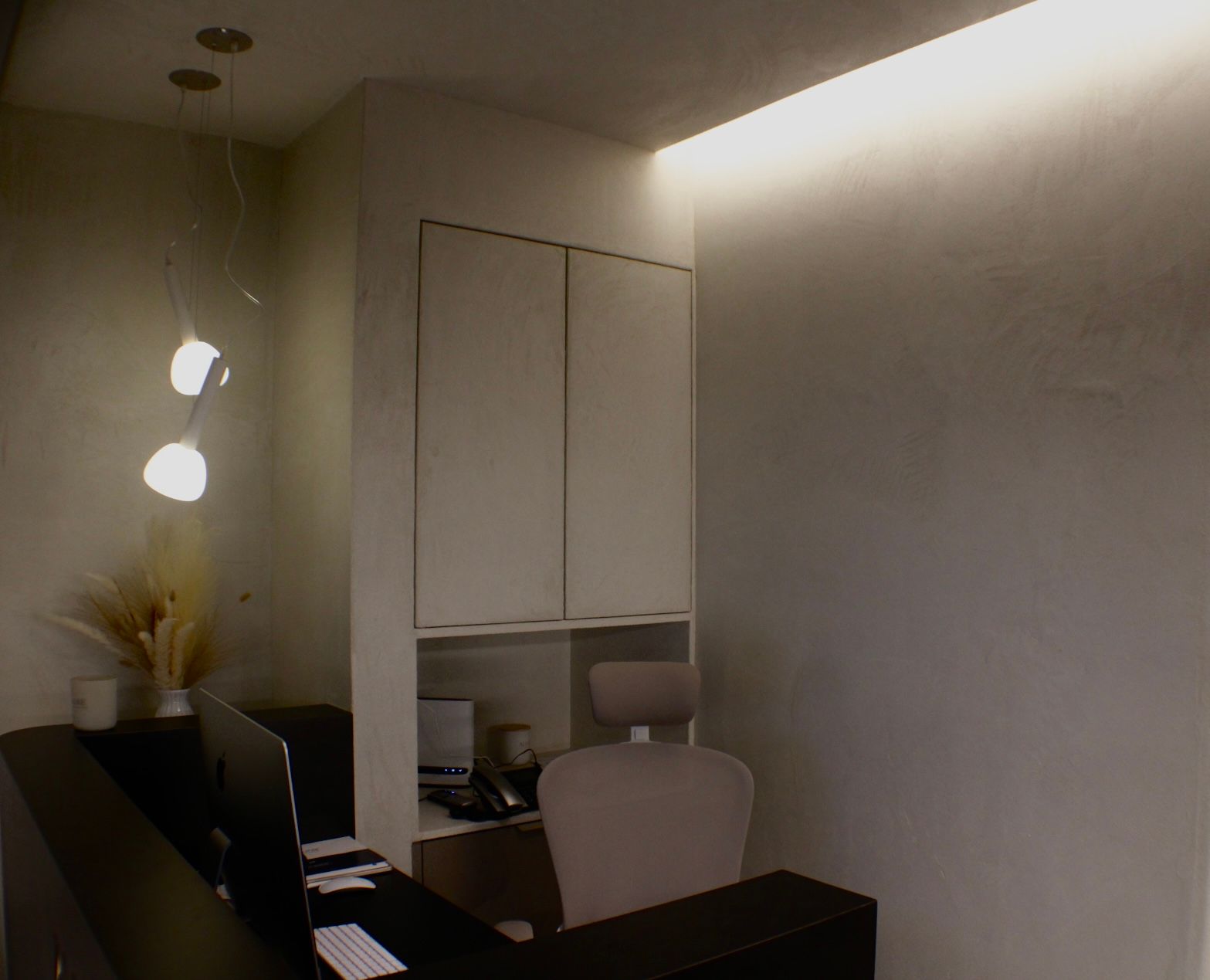 Espace réception : comptoir sombre, armoires, chaise de bureau, éclairage zénithal. Tons neutres, design moderne.