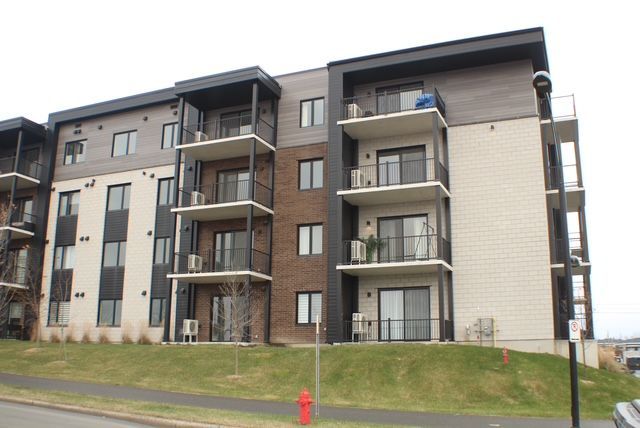 Immeuble d'appartements à plusieurs étages avec balcons; extérieur beige, marron et noir; sur une colline herbeuse.