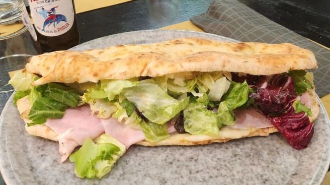 Panino con prosciutto e lattuga su un piatto di pietra, con una bottiglia di bevanda sullo sfondo.