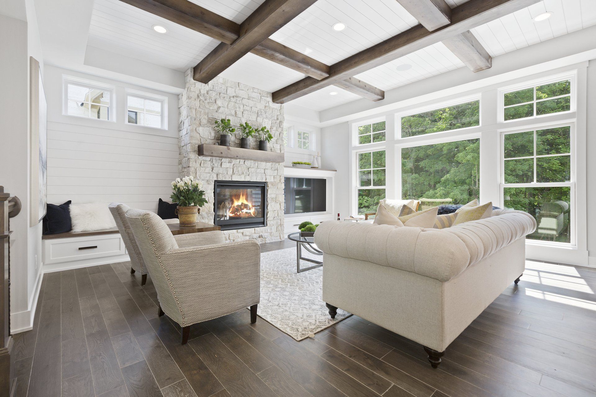 Living room with fireplace, white walls, wooden beams, large windows, and beige furniture.