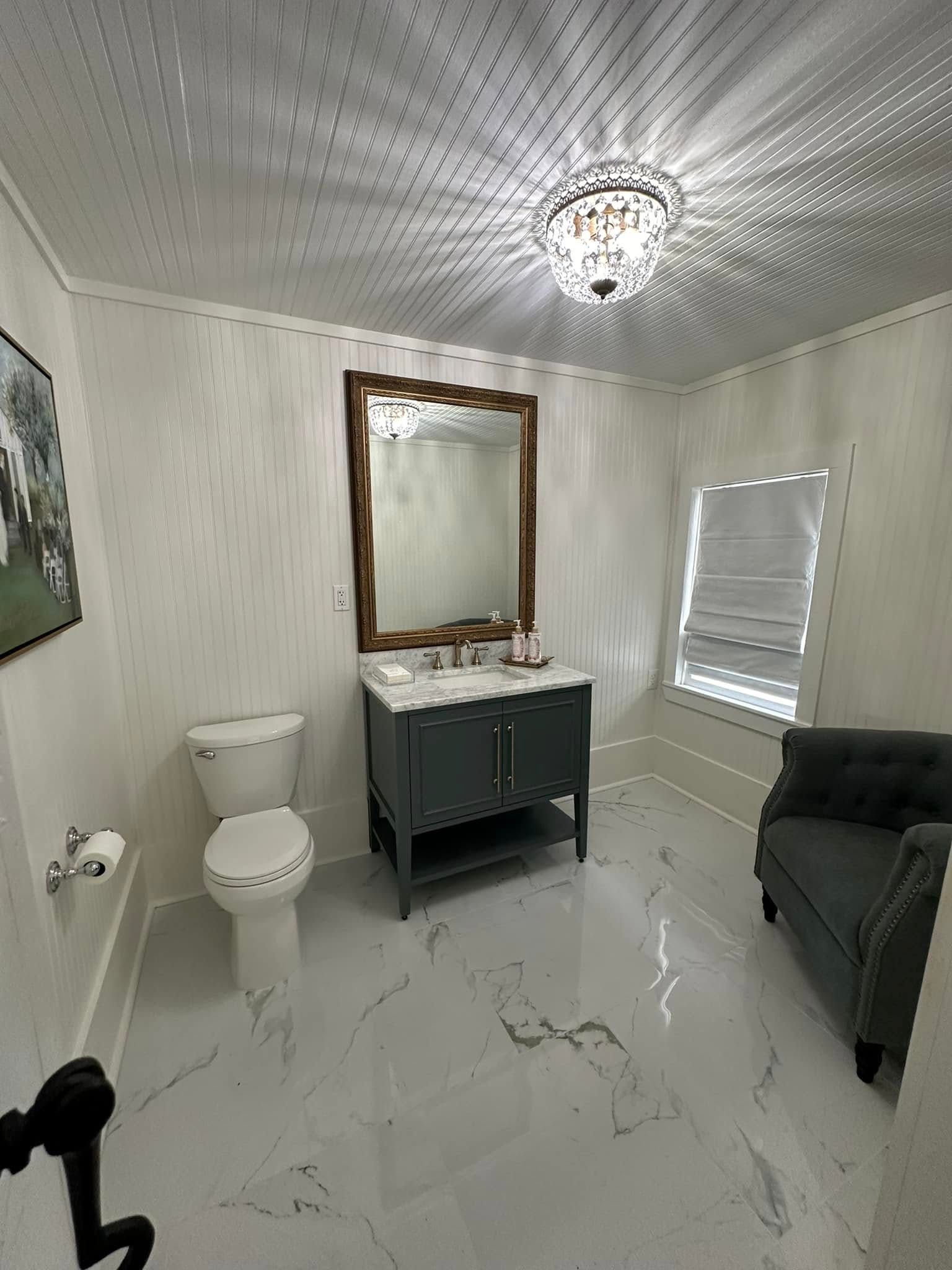 A bathroom with a toilet , sink , mirror and chair.