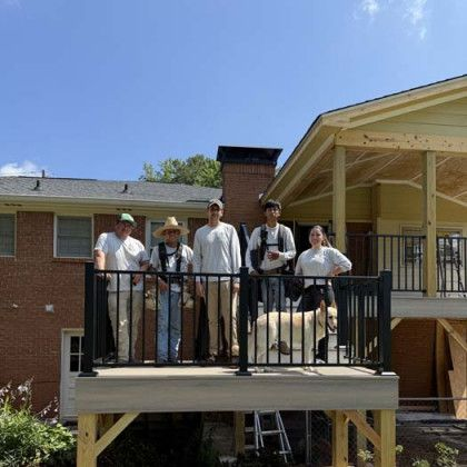 Five people standing on a balcony in front of a brick building under a blue sky