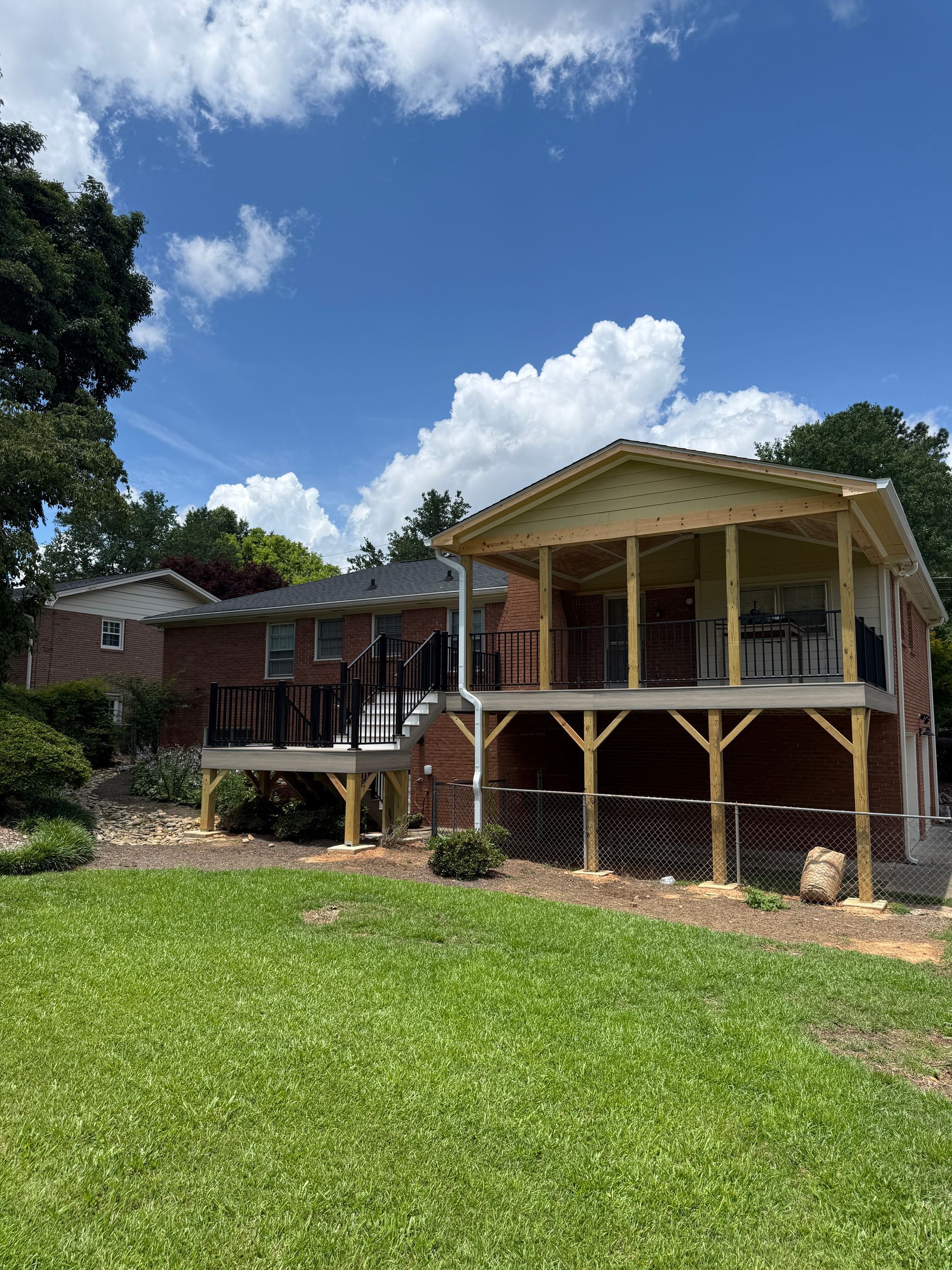 A brick house features a newly built, elevated wooden deck and covered porch overlooking a grassy lawn under a blue sky.