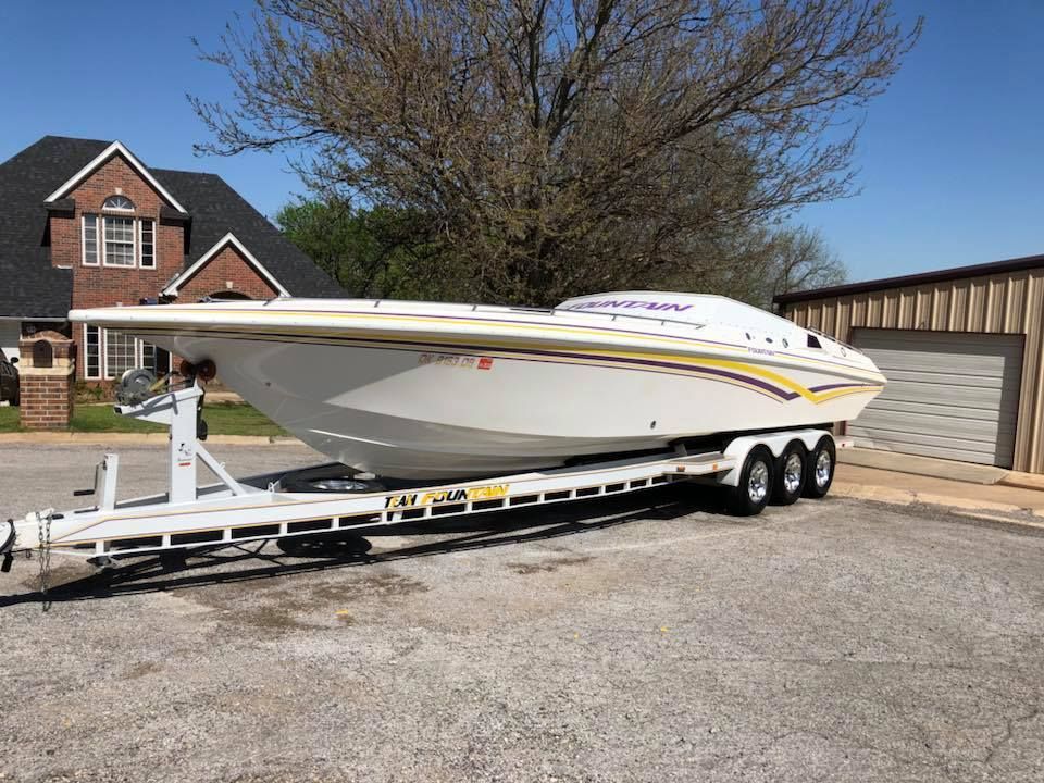 White speedboat on a trailer parked in a lot beside a building and house under a bare tree.