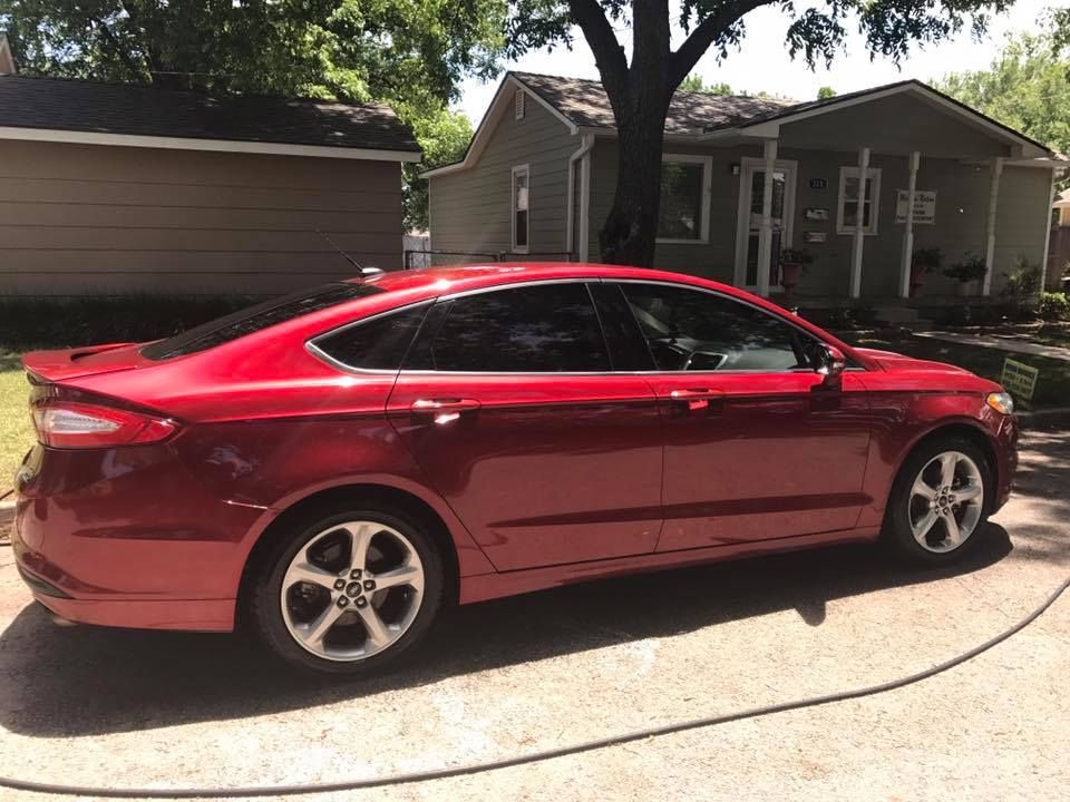 Red sedan parked in a driveway in front of a house, with trees and a garage in the background.