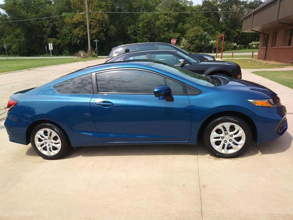 Blue coupe parked on a driveway with a black car behind it and trees in the background