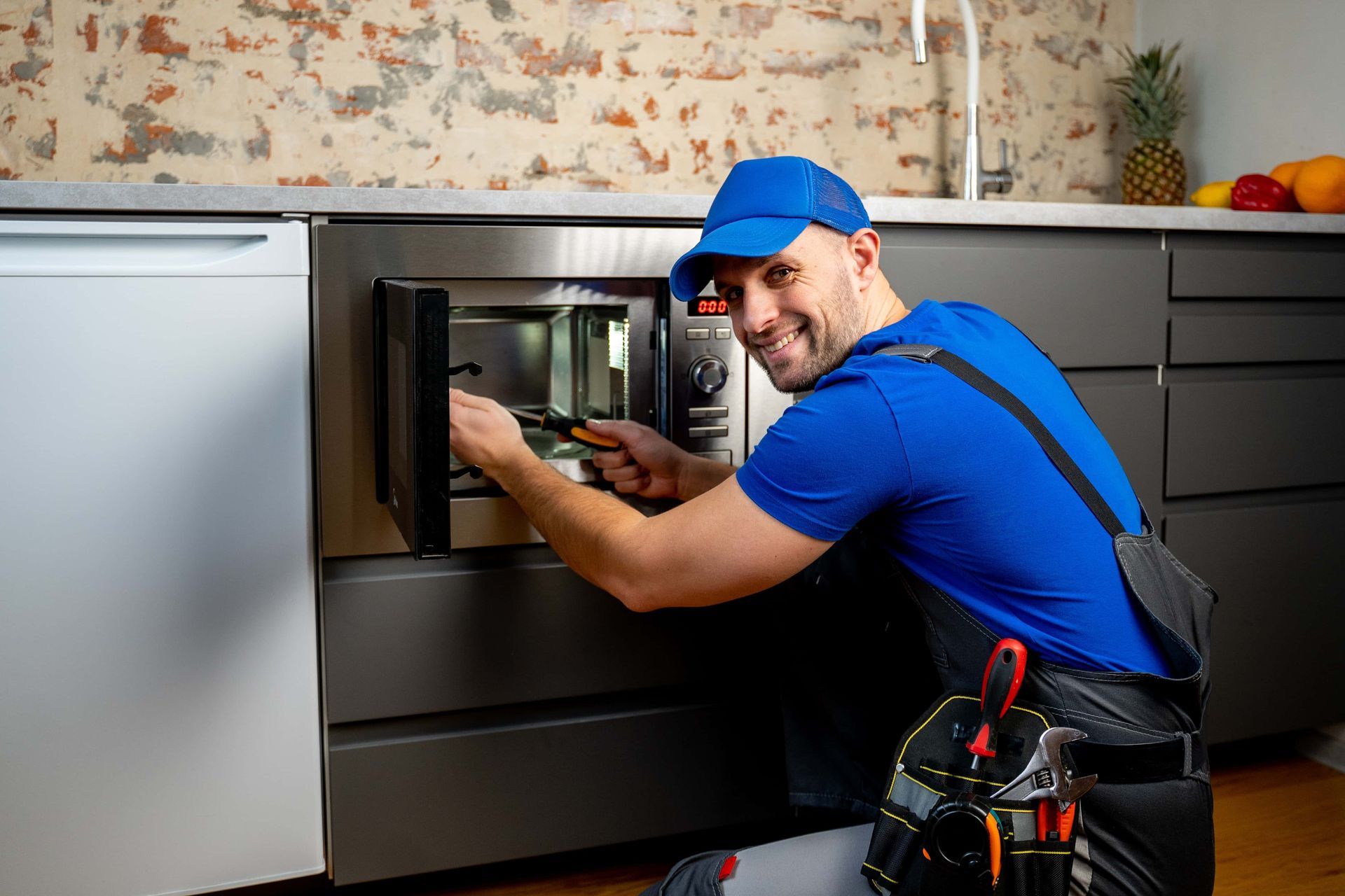Appliance technician repairing a built-in microwave in a modern kitchen, wearing a blue uniform and cap.