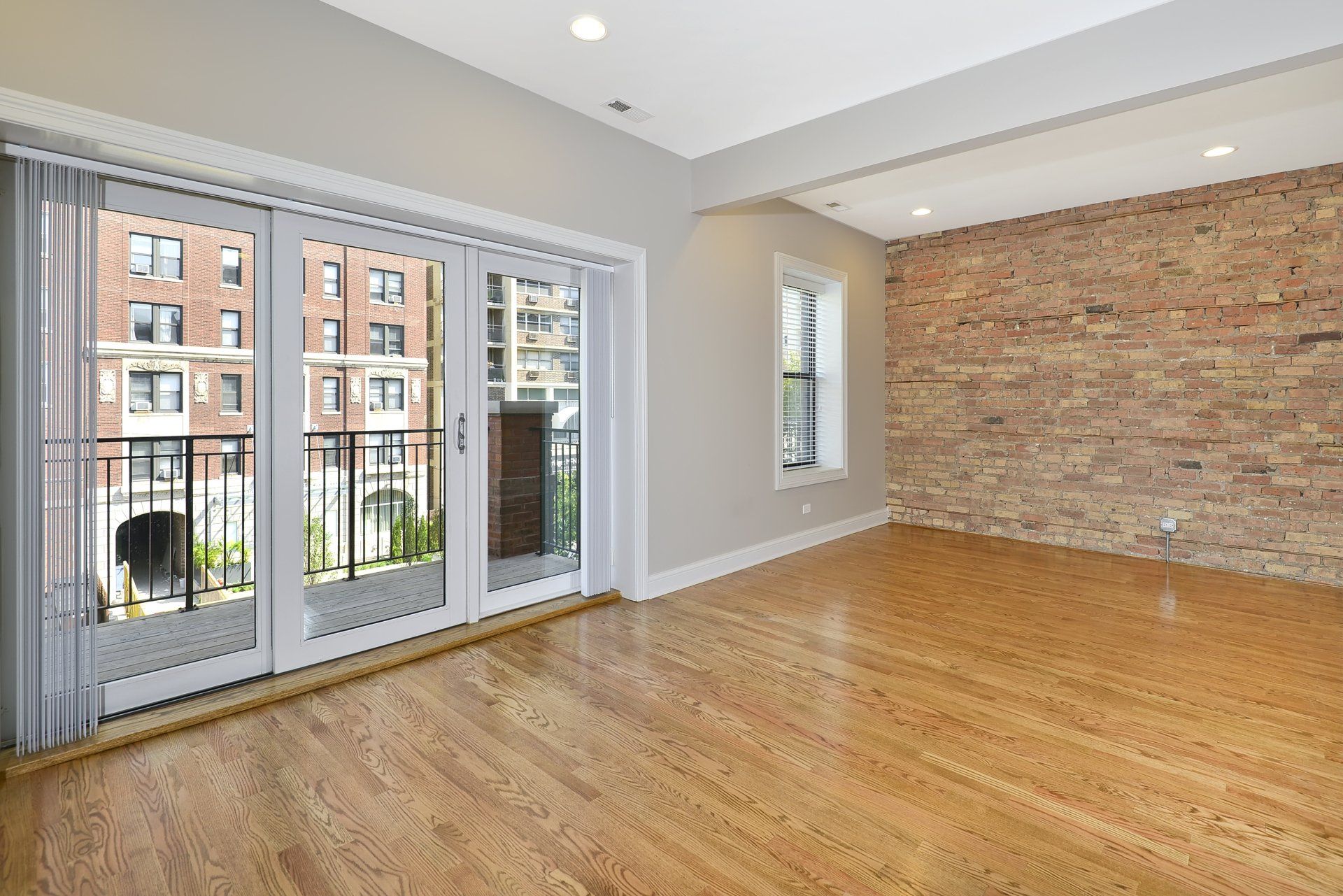 An empty living room with hardwood floors and a brick wall with exit door to the balcony.