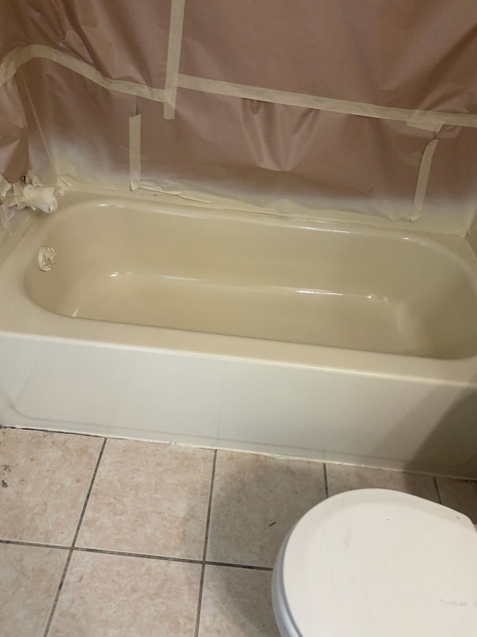 Bathroom bathtub with beige surround and protective plastic sheeting; toilet seat visible in foreground.