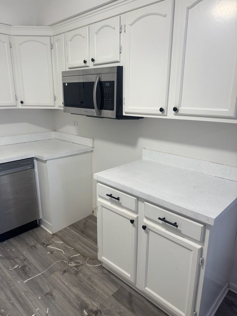 White kitchen with cabinets, black microwave, speckled countertops, and gray wood-look floor.