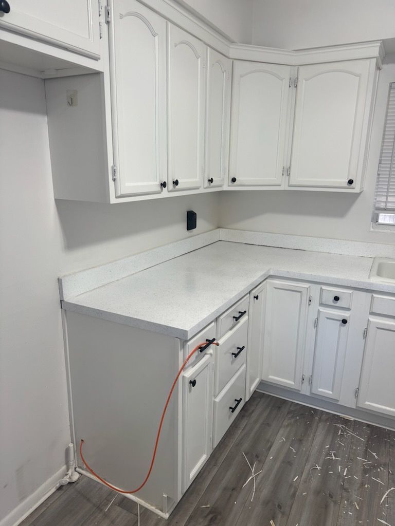 White kitchen corner with cabinets and countertops, unfinished with exposed dishwasher hose on gray floor.