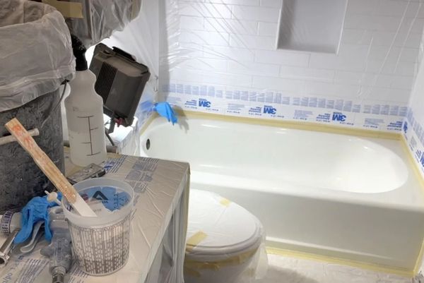 Painter in protective gear coating a bathroom tub with white paint, blue tape and supplies nearby