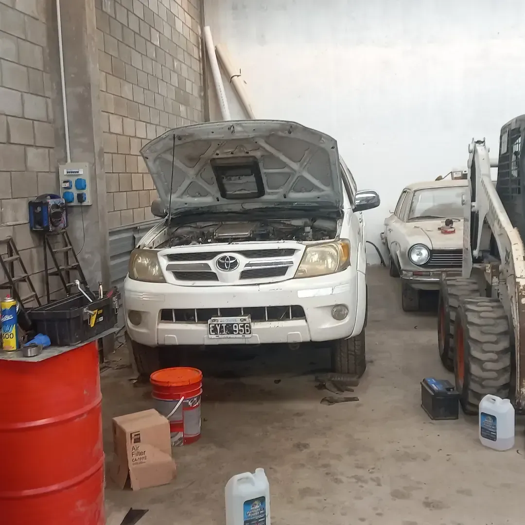 Una camioneta blanca con el capó abierto en un taller, estacionada junto a un auto antiguo y maquinaria de construcción.