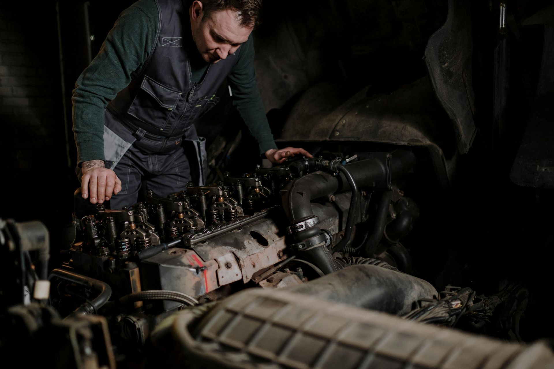 Un mecánico con ropa de trabajo oscura inspecciona el motor expuesto de un vehículo en un garaje con poca luz.