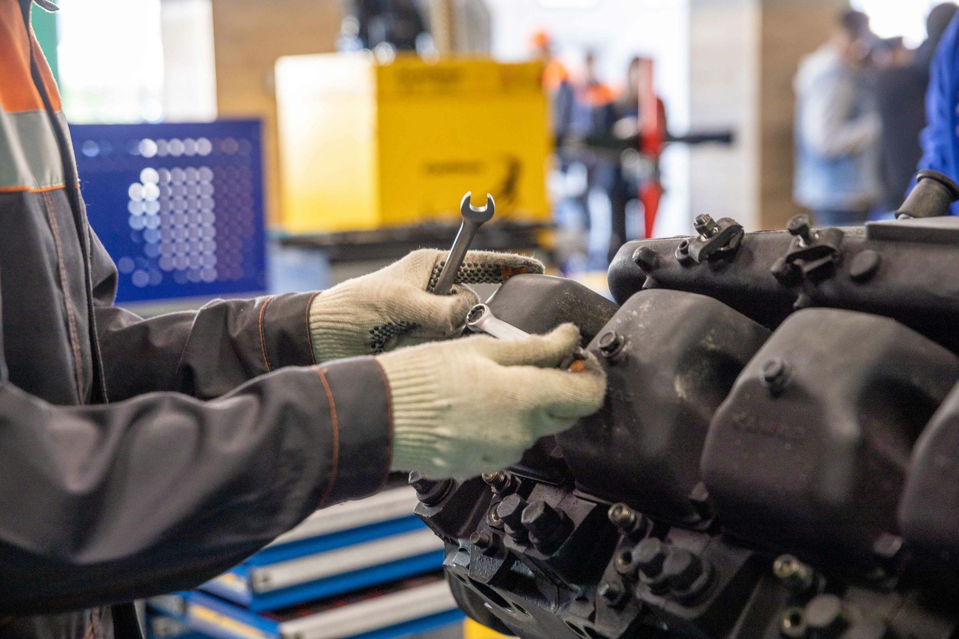 Una persona con mono de trabajo y guantes utiliza una llave inglesa para reparar un motor negro de gran tamaño en un taller.