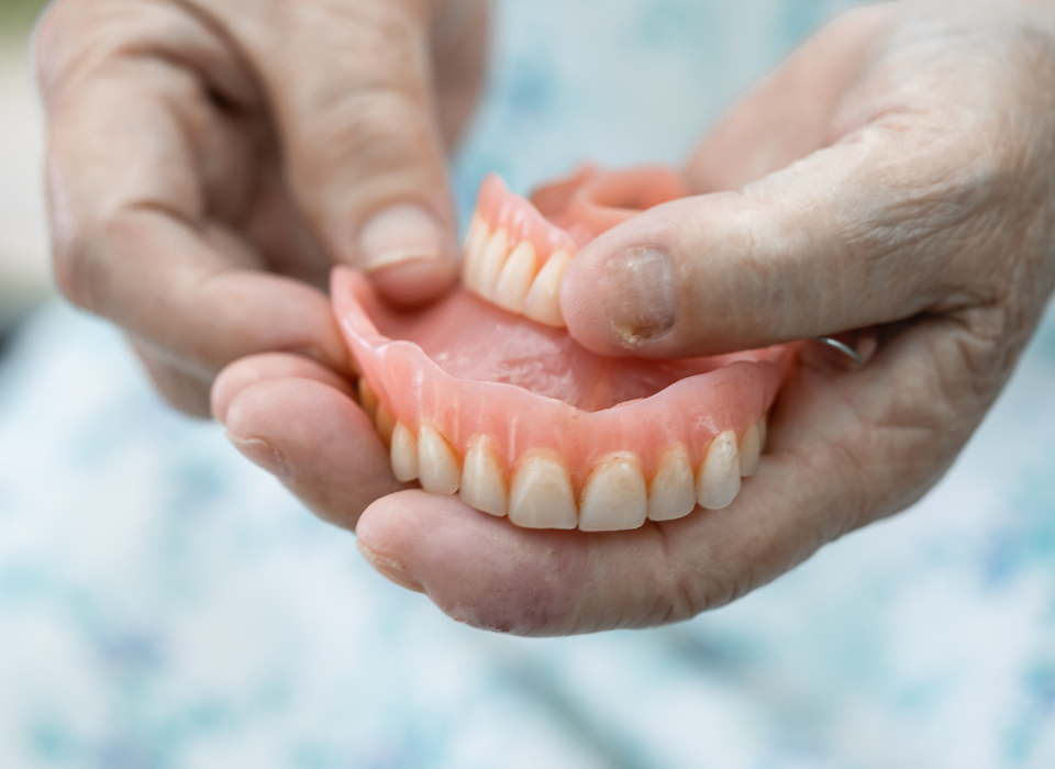 Hands holding a set of dentures. Pink gums and white teeth are visible.