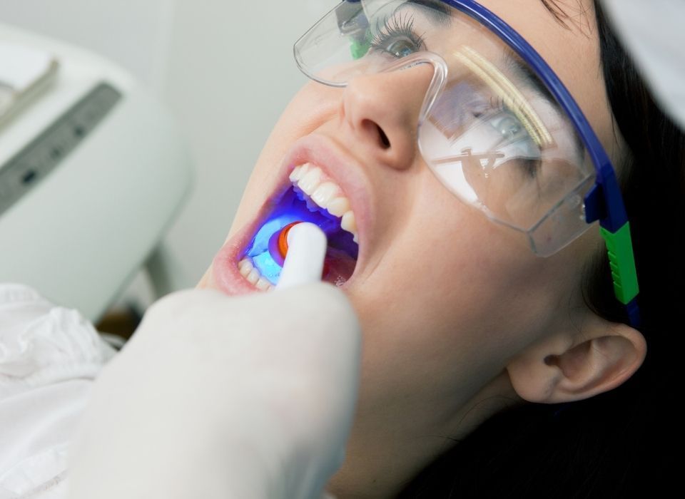 Dentist using a blue light on a patient's teeth; patient wearing protective glasses, mouth open, indoors.
