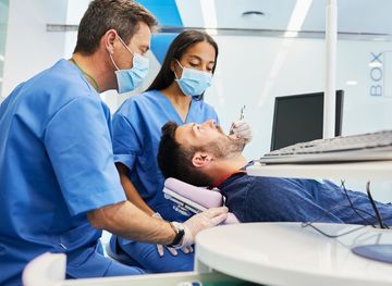 Woman in dental chair, holding cheek, looking concerned, presumably in pain, at dentist.