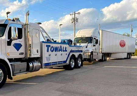 A tow truck is towing a semi truck in a parking lot.