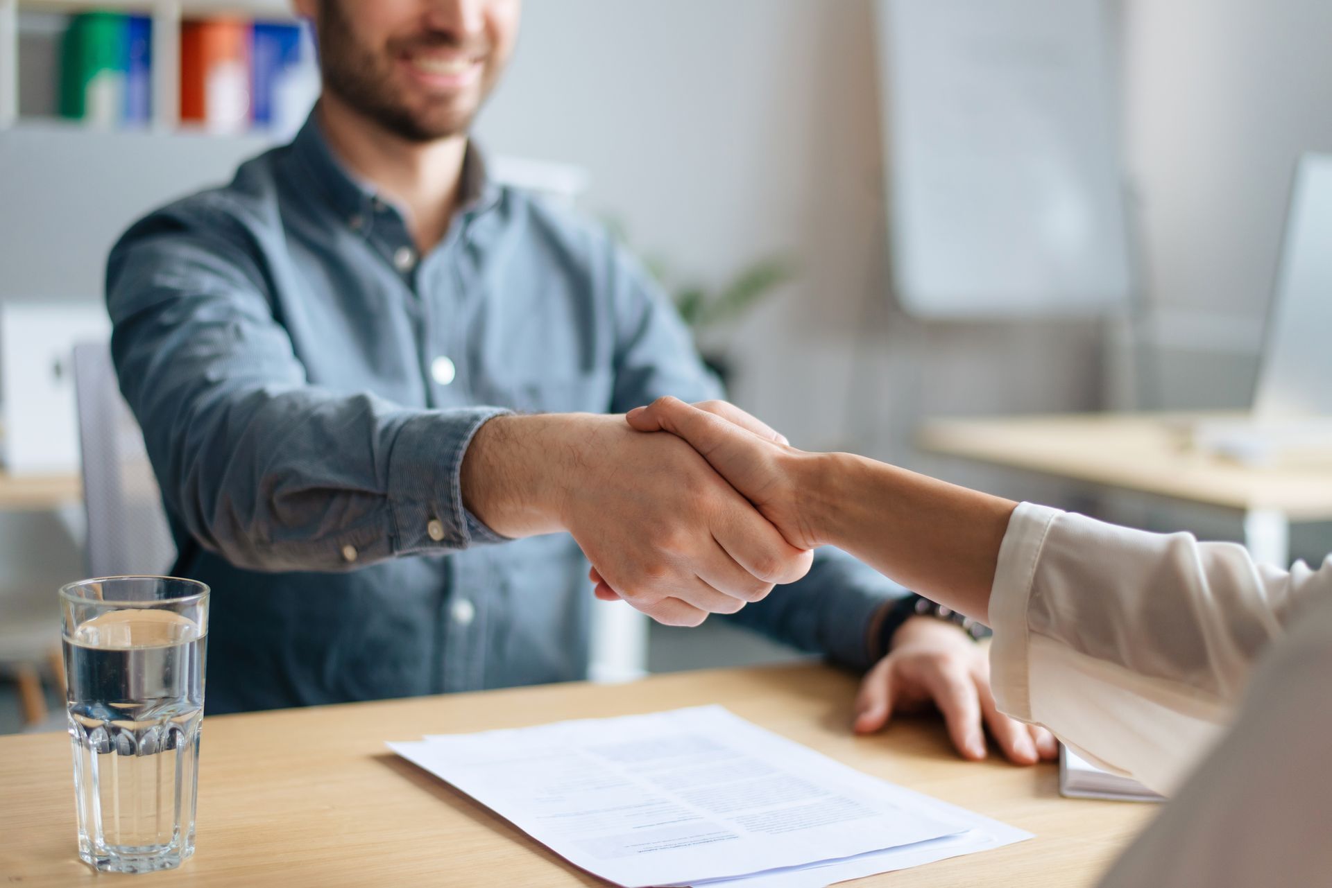 Two people shaking hands across a desk with a document and a glass of water nearby. Two people shaking hands across a desk with a document and a glass of water nearby.