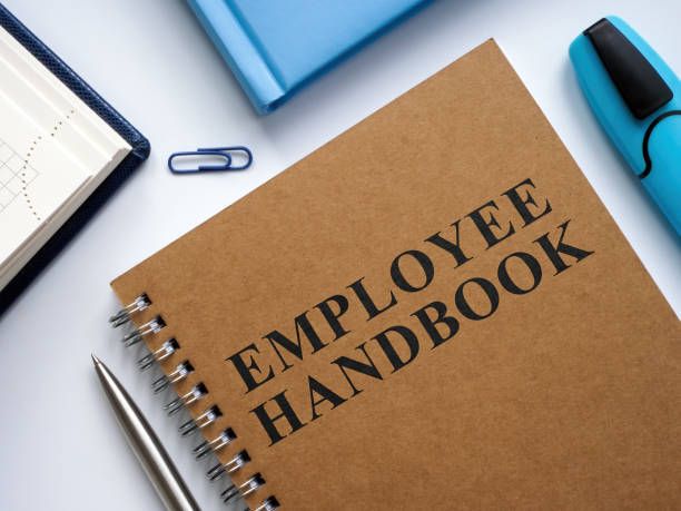 A spiral-bound notebook labeled 'Employee Handbook' lies among office supplies.