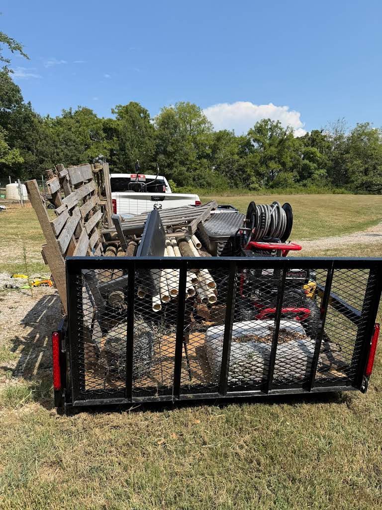 A utility trailer with firewood and equipment parked in a grassy field on a sunny day.