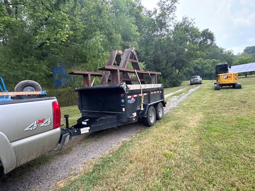 A truck pulling a trailer with a metal structure on a grassy path, with vehicles and trees in the background.