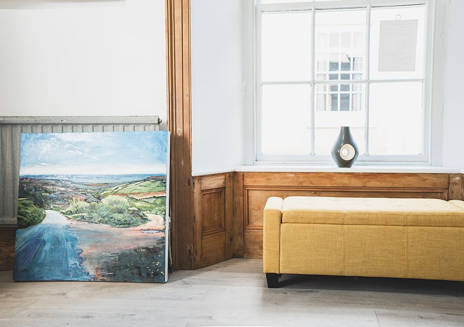 inside gallery, light grey floor, white walls and ceiling. There are two large window reveals framed with pine architrave, the large skirting board is also pine. There are three paintings hanging, they look like sea scapes and have white frames. There are cushions stacked in the corner and a yellow bench seat in the window. There is shelving with lots of tubes of paint. 