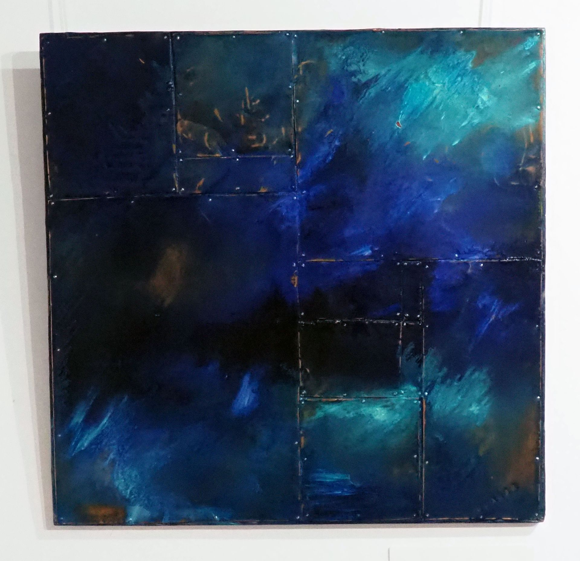 Large and small rectangles of copper pinned onto a copper coloured board. The copper is tarnished in places. Blues are painted with aqua over the piece. The aqua and some of the blue look iridescent.