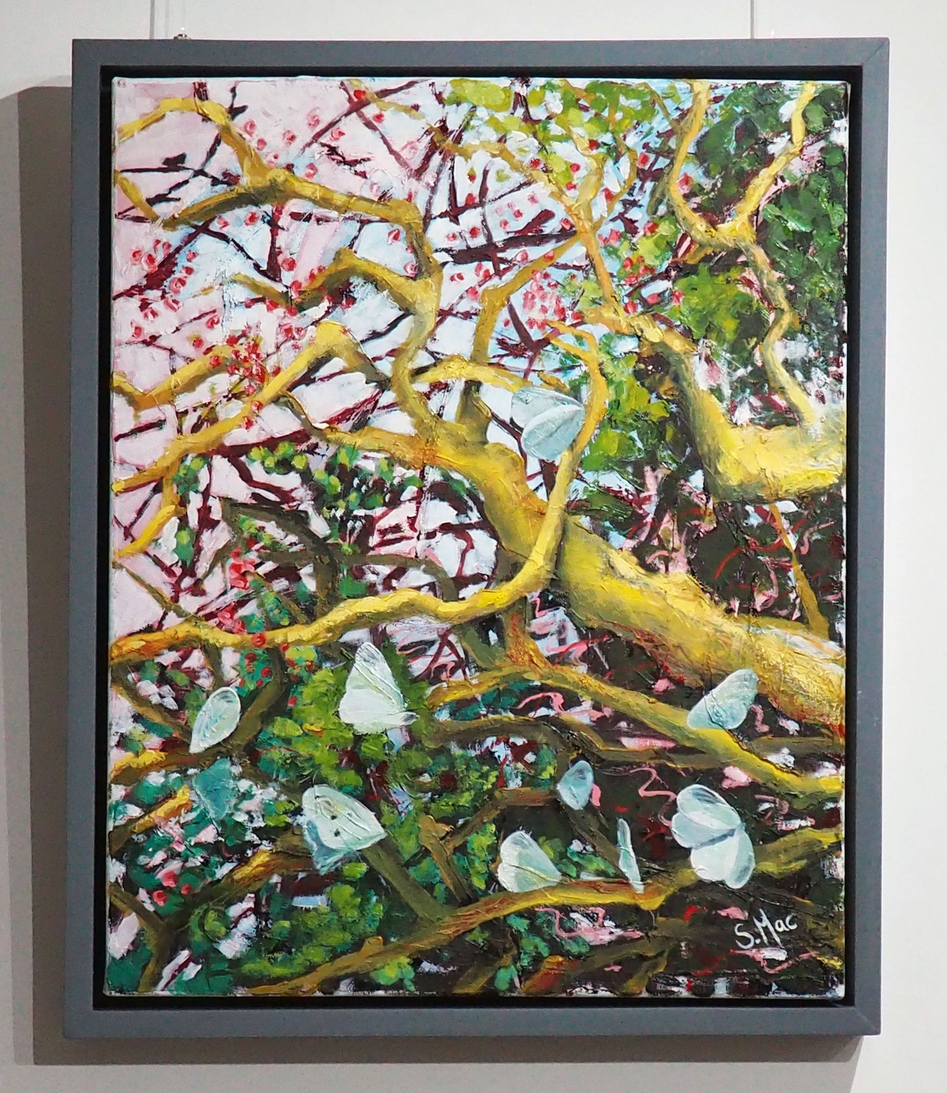 A portrait orientated painting of branches with around a dozen pale grey / green butterflies. The branches in the foreground are an ochre glow. Towards the bottom half and top right there is forest and acid green foliage, and the butterflies are place in various positions of wings open and closed. In the top left there is light blue and light pink sky showing red pink berries on the bare branches. Framed in a simple grey wood.