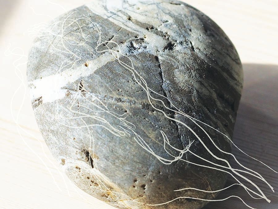 A photographic close up of a grey pebble with white marks on it. Imposed on the pebble is simple grey line drawings suggesting a naked person laid across the pebble.