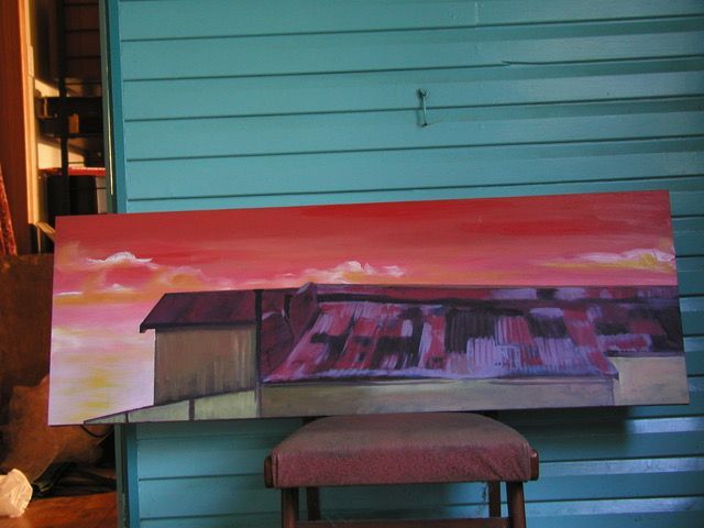 A photo of a long landscape painting perched on a chair. The paintings is of a huge corrugated roof. The colours of the roof are cold reds and purples. The building is set against a red sunsetting sky with a few wispy orange and white clouds