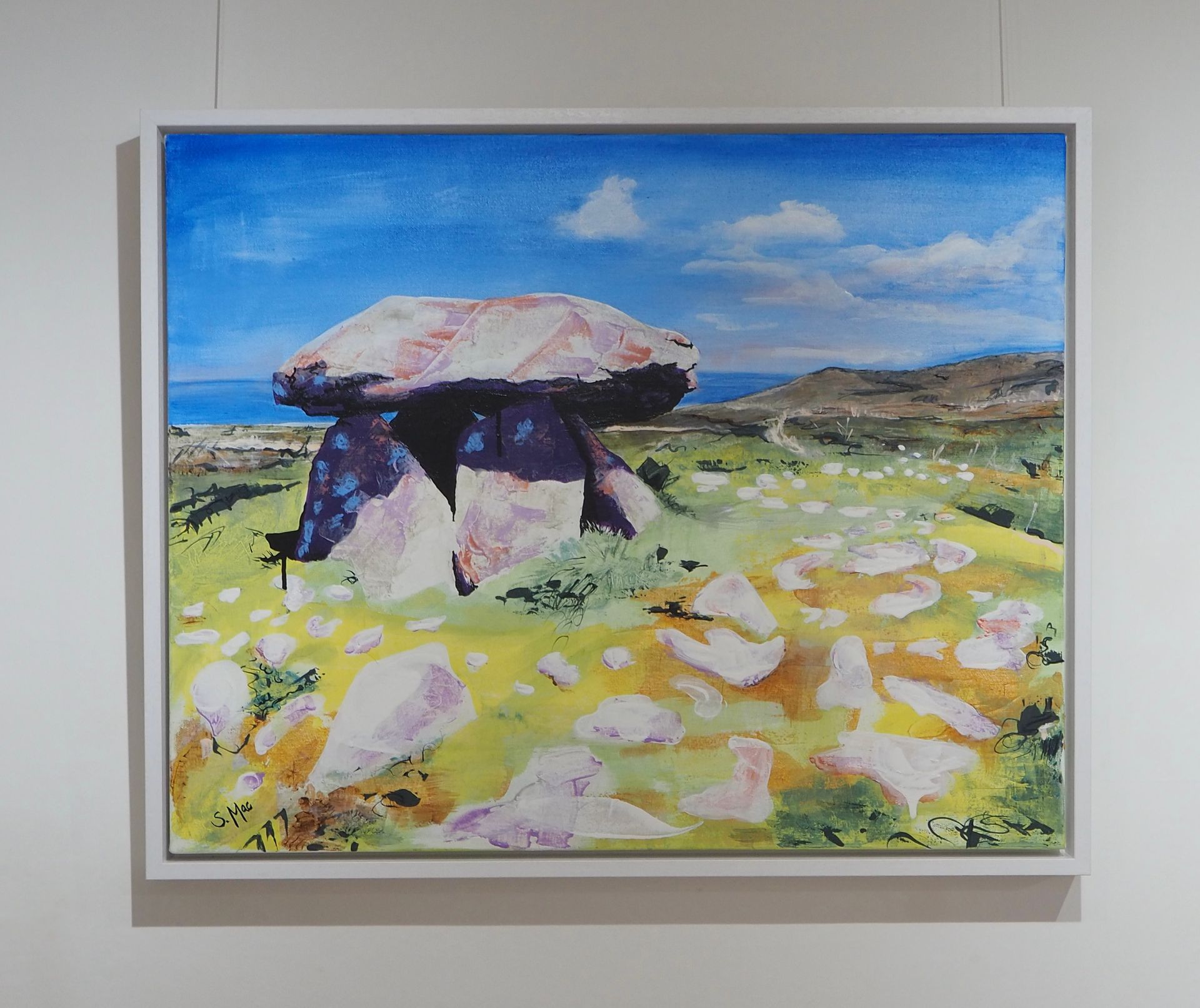 A stone structure on a landscape with blue sea and sky in the background. The ancient structure is detailed with contrasting shadows  to the sun drenched landscape. large rocks are in the foreground which is coloured yellow, orange and green. Darker hills are to the right on the horizon.