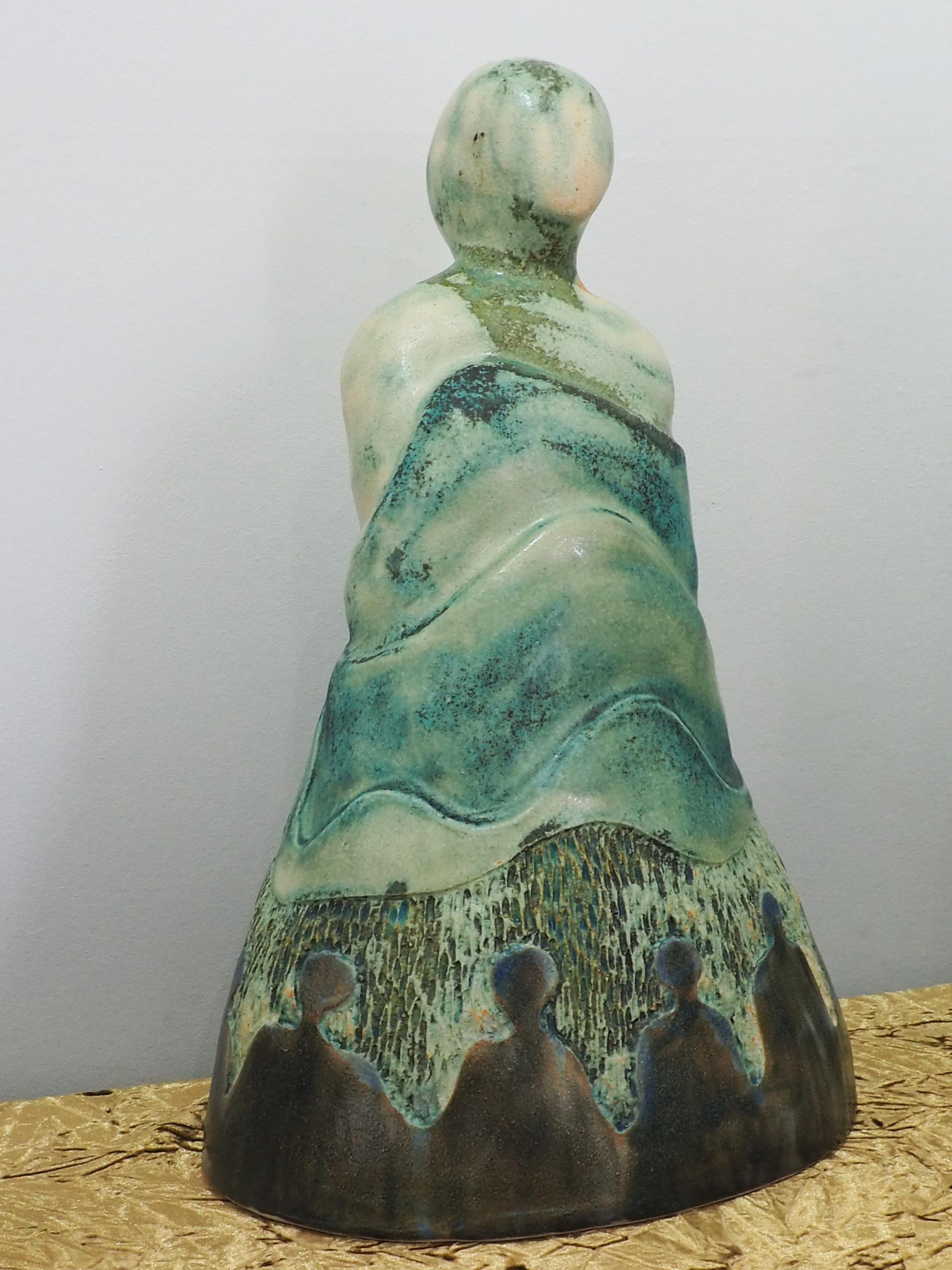 A sculpture of a smooth figure coming out of smooth ripling layers. The glaze is bronze brown at the bottom and greens towards the top. Skirting the bottom are figures of people against a rough texture