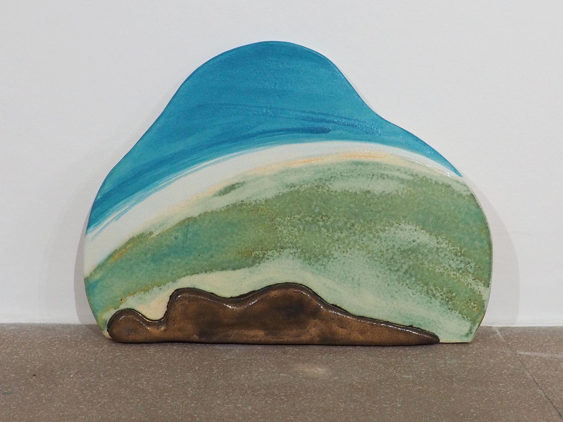 A vertical slab sculpture with a silhouetted figure reclined in the foreground. The figure is glazed bronzy brown and the slab background is muted greens, then an off white to a aqua blue at the top. Brush marks are shown in these colours.