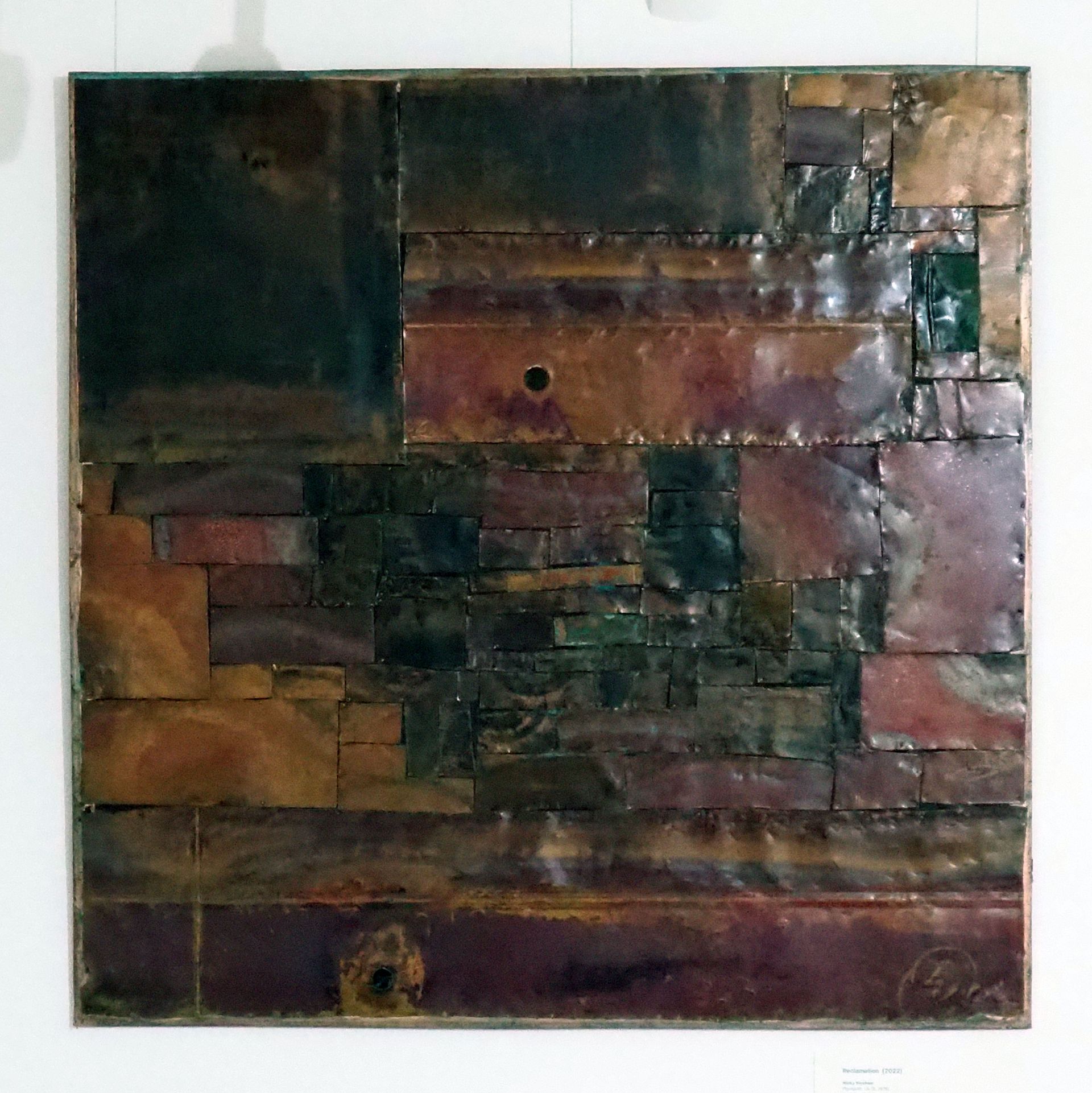 Around sixty large and small rectangles of copper pinned together to form a square. The rectangles of copper are tarnished with different colours - black / green and maroon purple and copper. 