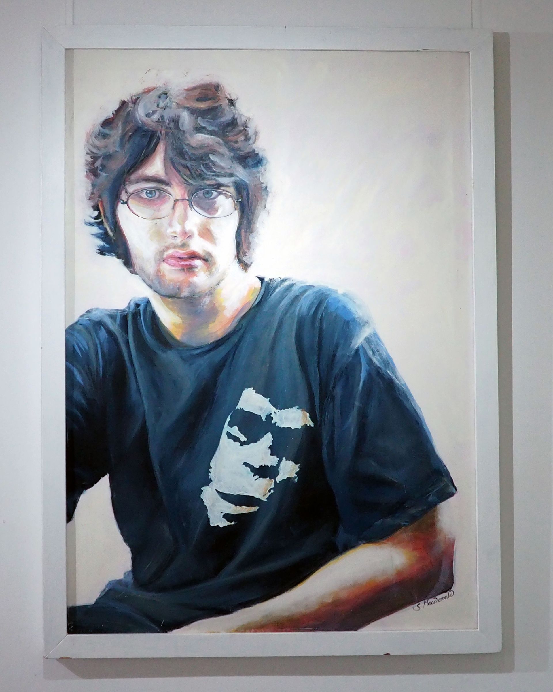 A large portrait of a young man wearing a black T-shirt with a white stencil of a gorilla's face on it. The man has shaggy hair and stares straight forward at the viewer. The background is white, the subject is saturated in this white 'light'.