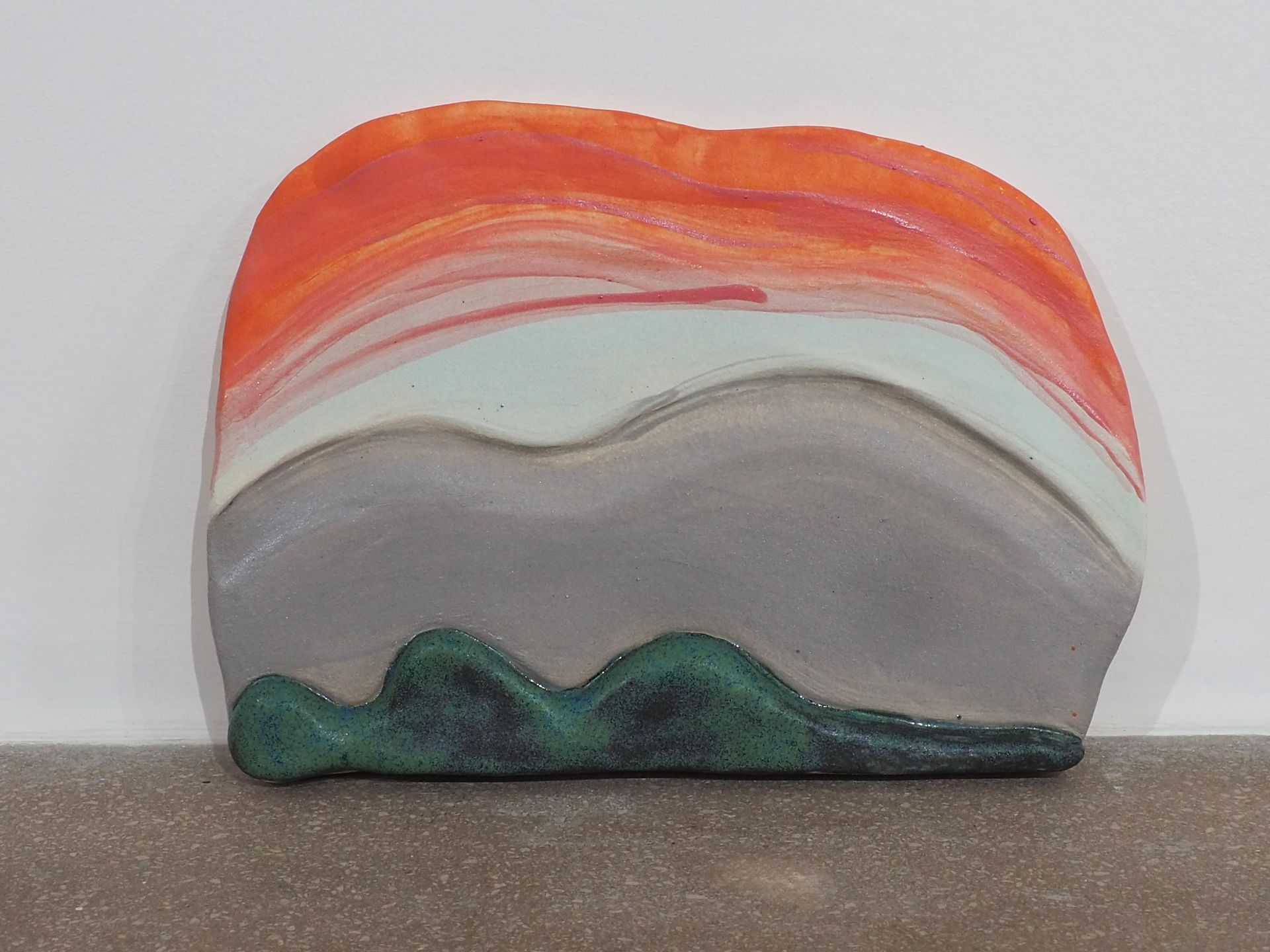 A vertical slab sculpture with a silhouetted figure reclined in the foreground. The figure is glazed dark greens and the slab background is a lavender grey, then an off white to an orange red at the top. Brush marks are shown in these colours.