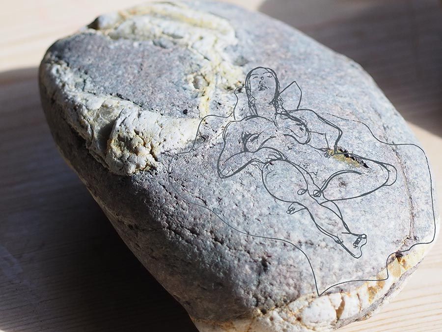 A photographic close up of a grey pebble with white marks on it. Imposed on the pebble is a simple grey line drawing of a naked woman lying down on her back. 