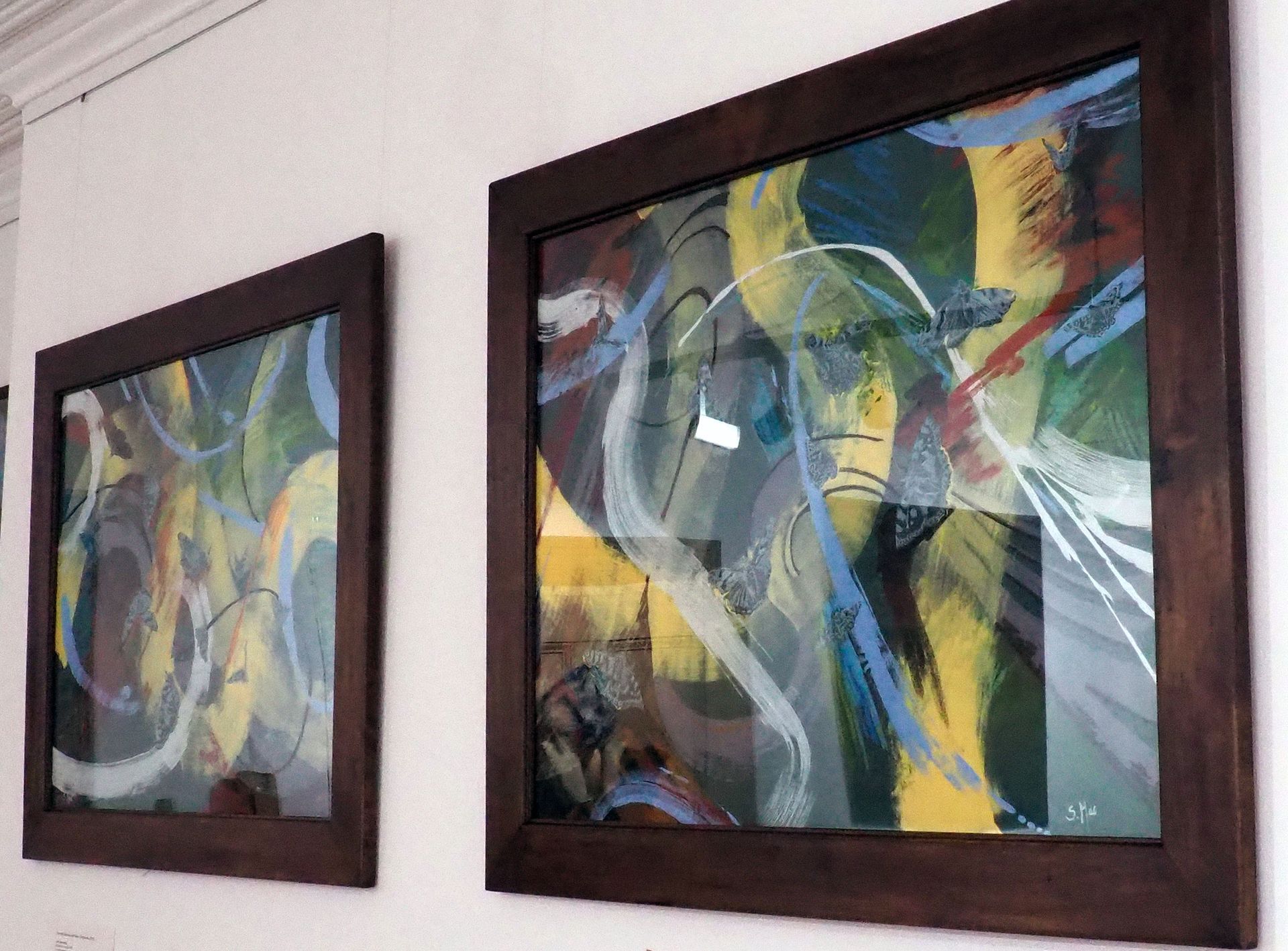 A photo of two large landscape dark wood frames taken at an angle. The framed images are behind perspex and there is a reflection. The images consist of large yellow, white and blue brush trails that flow from one frame to the other. The pieces belong together as a pair as they are 'joined' by the largest yellow brush trail . The background is dark green and there are around 14 delicate grey butterflies that move amongst the brush marks.