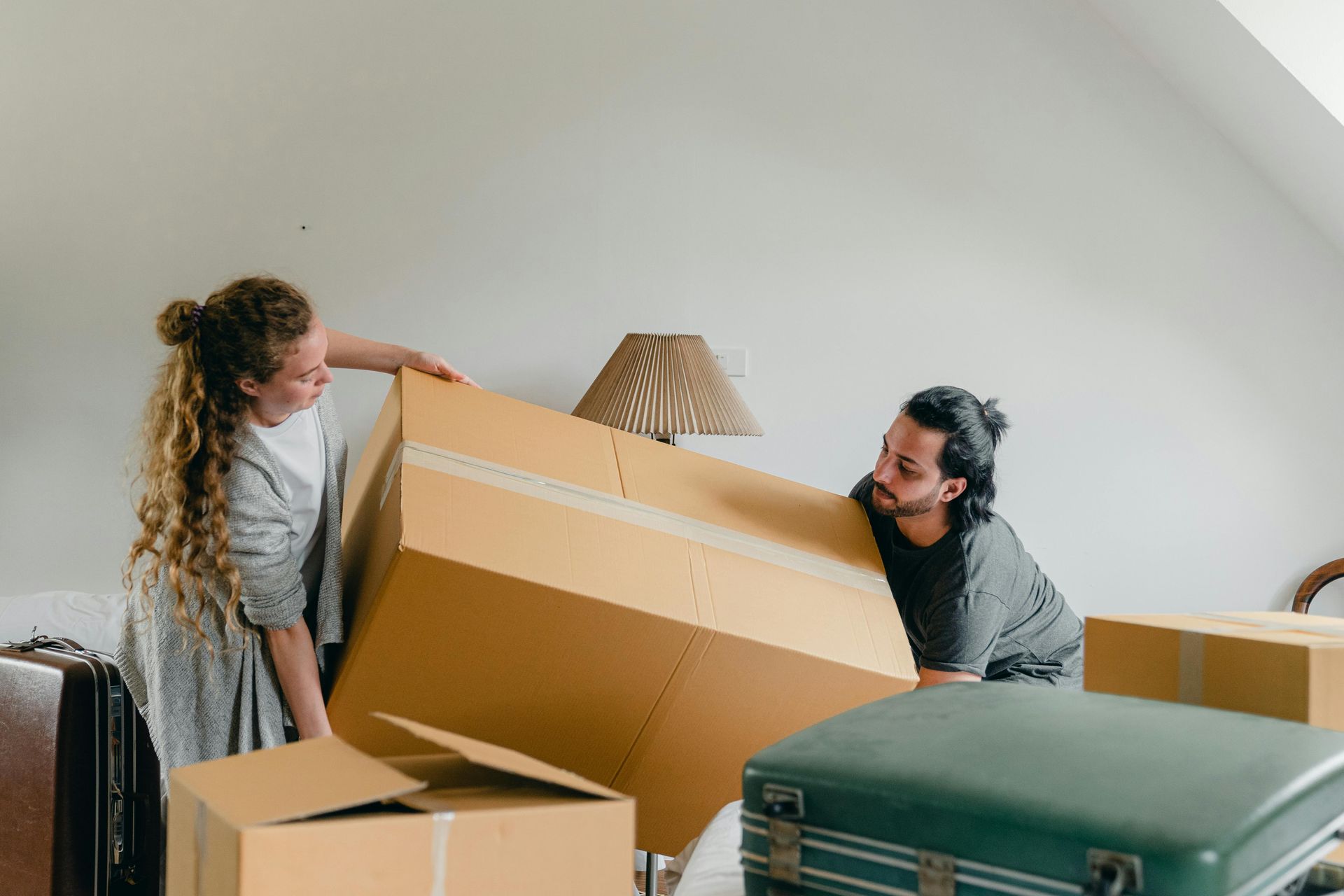Couple lifting a large cardboard box in a room with other boxes, possibly moving.