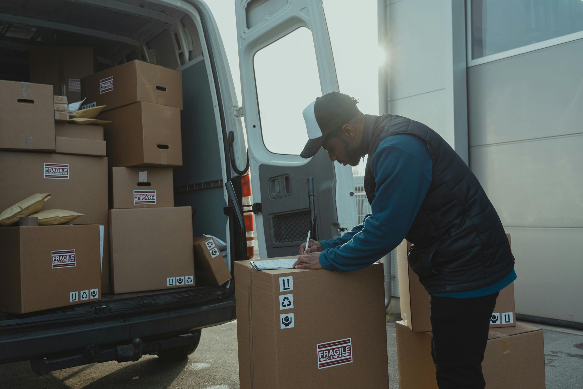 Delivery person writing on a package next to a van full of boxes in daylight.