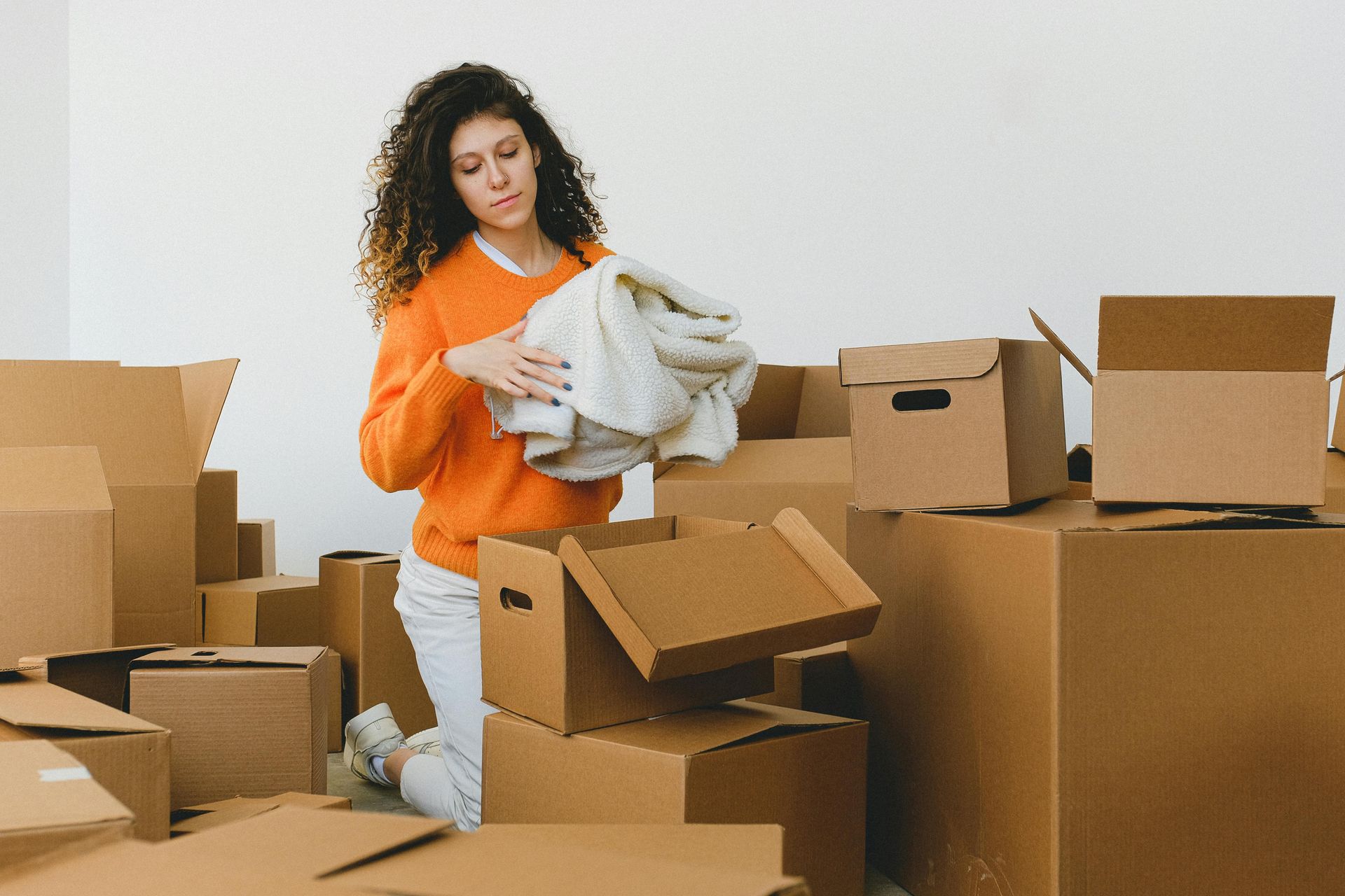 Woman in orange sweater packing clothes into cardboard boxes in a room.