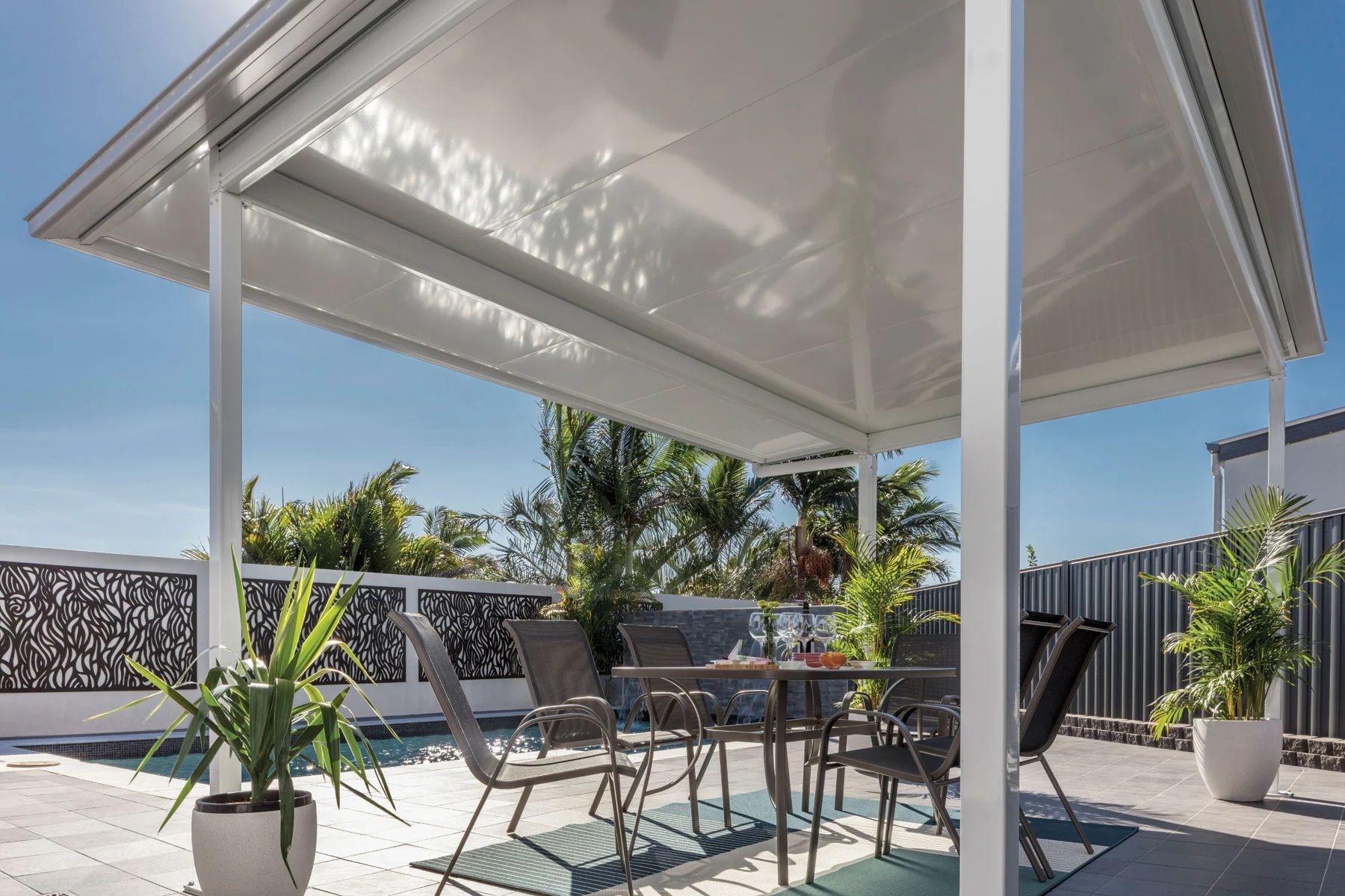 Outdoor patio with white pergola, glass table, black chairs, and potted plants under a blue sky