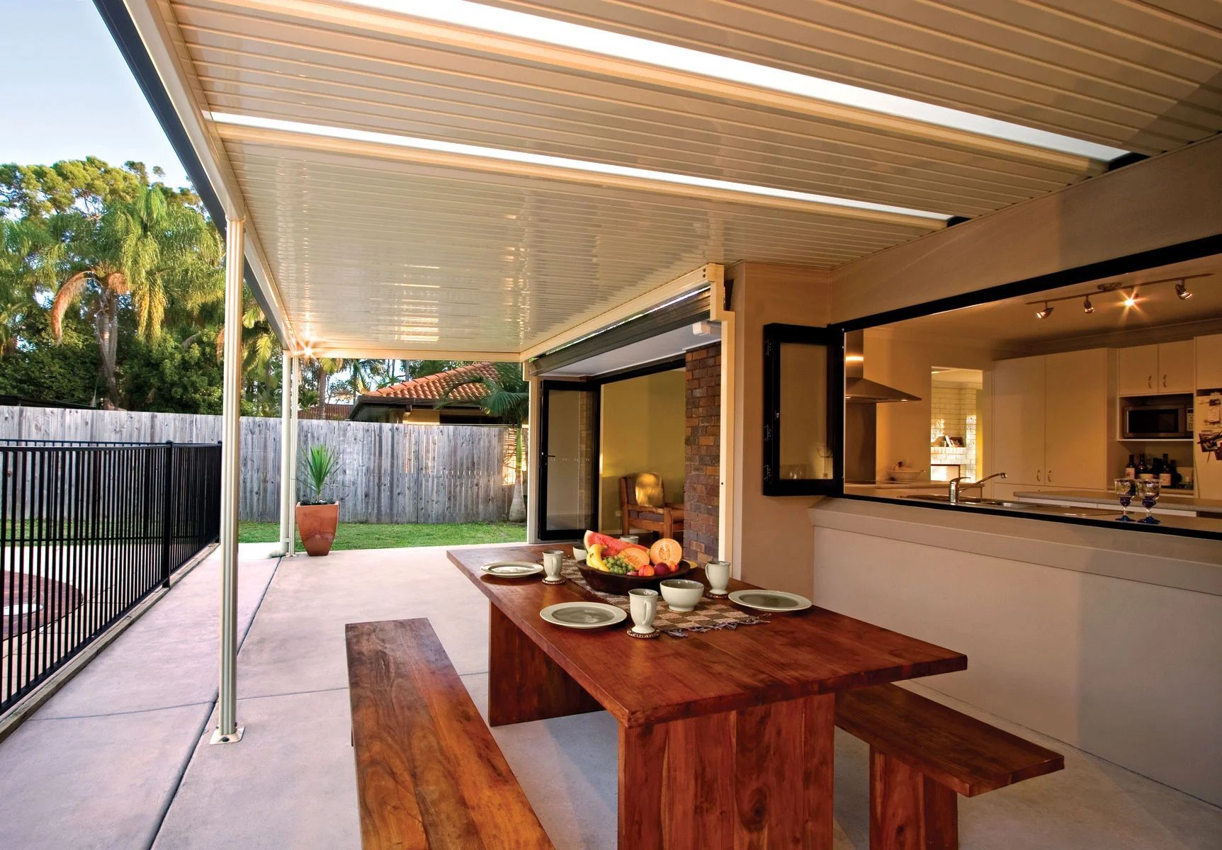Covered patio with wooden picnic table, view of indoor kitchen, and fenced backyard beyond.