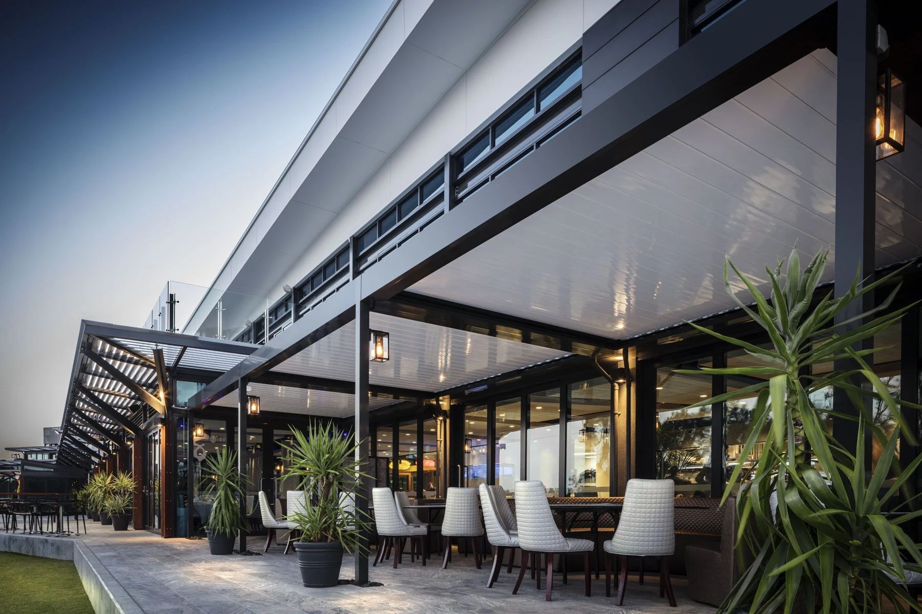 Modern outdoor restaurant patio with white chairs, black tables, potted plants, and a long canopy