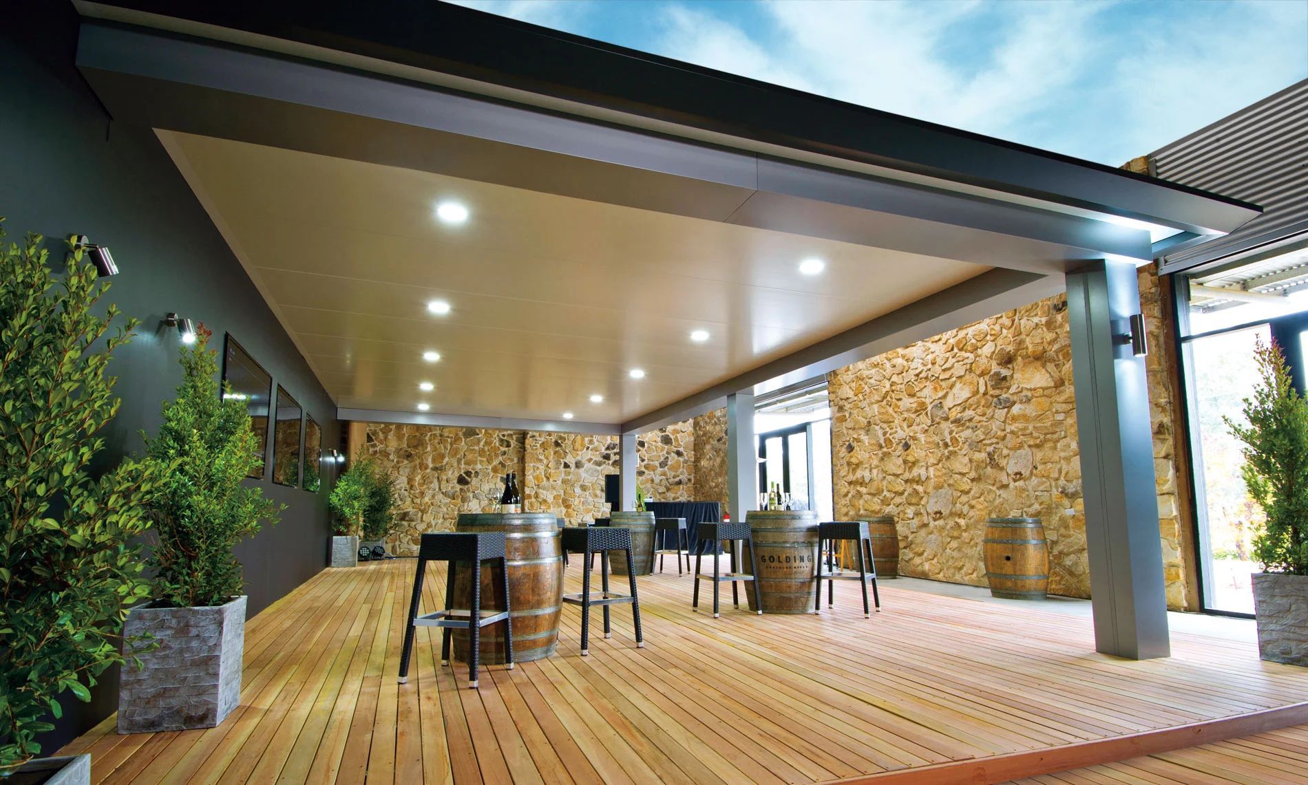 Modern covered patio with wooden deck, bar stools, stone wall, and recessed ceiling lights