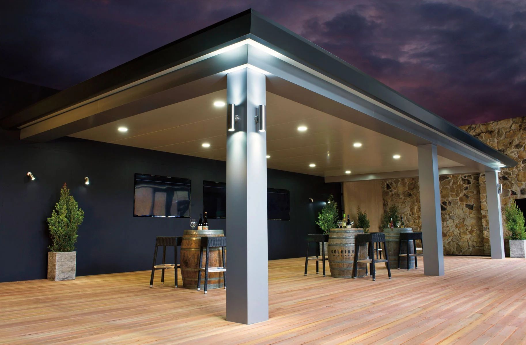 Modern outdoor patio with wooden deck, bar stools, tables, and a lit covered roof at dusk